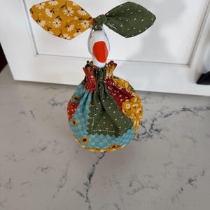 May include: A decorative goose figurine featuring a white head with an orange beak, adorned with a fabric bow on its head and a patchwork fabric cover over its body. The fabric features various patterns including sunflowers, small white flowers, and green dots.
