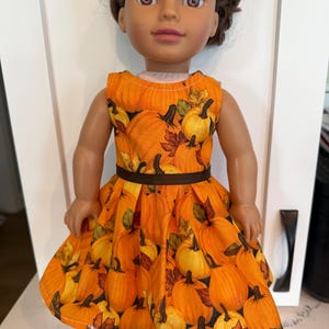 May include: A doll with brown hair and brown eyes wearing a sleeveless dress featuring an orange and yellow pumpkin and autumn leaf print. The dress has a fitted bodice, a dark brown ribbon waistband, and a pleated skirt that flares outward.