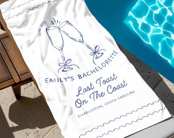 May include: White beach towel with blue text and a graphic of two champagne glasses clinking. The text reads "Emily's Bachelorette Last Toast On The Coast Charleston, South Carolina June 2023".