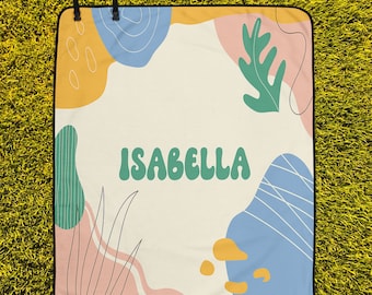 May include: A white picnic blanket with a black border. The blanket has a colorful abstract design with green leaves, yellow and blue shapes, and the name "ISABELLA" in green letters.