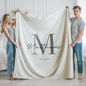 May include: A white fleece blanket with a large black letter "M" and the text "Matthews EST 2024" printed on it. Two people are holding the blanket up.