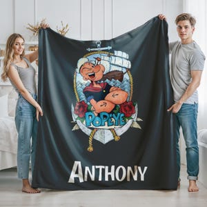 May include: A black blanket featuring a cartoon image of Popeye the Sailor Man inside an anchor design, with the name "Anthony" in white block letters at the bottom. The blanket has a nautical theme with red roses and rope accents.
