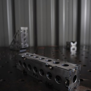 Könnte beinhalten: Dunkler rechteckiger Metallblock mit mehreren kreisförmigen Löchern und Ausschnittdetails. Der Block befindet sich auf einer dunklen Oberfläche mit anderen Metallteilen im Hintergrund. Das Industriedesign deutet auf eine Werkstatt oder eine Fertigungsumgebung hin.