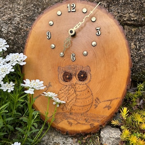 May include: A wooden clock with a natural edge, featuring an engraved owl design. The clock face has black numerals and gold-colored hands. The clock is approximately 10 inches in diameter and is set against a natural background with flowers.