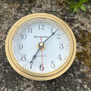 May include: A round, gold-toned Brookstone quartz clock with a white face. Black hands indicate the time, with the numbers 1 through 12 in black. The clock is set against a textured, gray surface, with green foliage in the background.