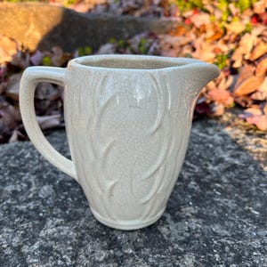 May include: A white ceramic pitcher with a crackled glaze and a leaf design. The pitcher has a handle and a spout.