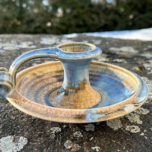 May include: Handmade ceramic candle holder with a handle. The holder features a circular base and a central cup for the candle. The color scheme includes shades of blue, brown, and tan, with a glossy finish. The handle is arched and attached to the side.