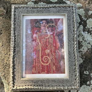 May include: A framed print featuring a woman in a red and gold dress, holding a snake. The artwork has a vintage aesthetic, with a detailed silver-colored frame. The print is set against a natural stone background.