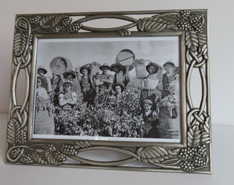 May include: A silver-toned picture frame with an ornate design featuring grapevines and berries. The frame holds a black and white photograph of a group of people harvesting grapes.