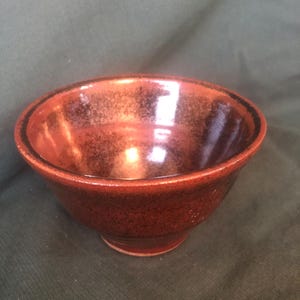 May include: A small, round ceramic bowl with a speckled, reddish-brown glaze. The bowl has a slightly flared rim and a small, rounded base. The interior reflects light, and the exterior has a textured appearance.
