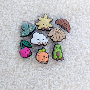 May include: A selection of colourful shoe charms with different designs. The charms include an octopus, sun, croissant, bunny, cloud, cherry, pumpkin, and avocado. Each charm has a smiling face and is made of a rubbery material.