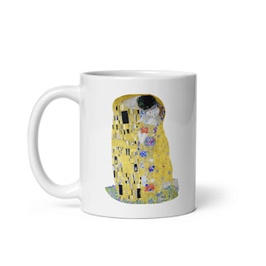 Puede incluir: Taza de cerámica blanca con una imagen colorida de la pintura "El Beso" de Gustav Klimt. La pintura representa dos figuras abrazándose en un patrón de mosaico dorado.