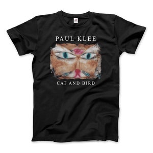 Puede incluir: Camiseta negra con una impresión blanca de una pintura de Paul Klee titulada "Gato y pájaro". La pintura presenta una cara de gato estilizada con ojos azules y una nariz rosa.