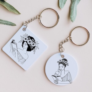 May include: Two white keychains with black line drawings of women reading books. Both women have floral crowns and are drawn in a minimalist style.
