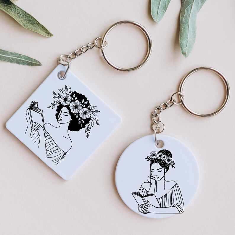 May include: Two white keychains with black line drawings of women reading books. Both women have floral crowns and are drawn in a minimalist style.