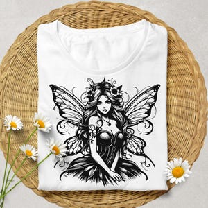 May include: White t-shirt featuring a black and white illustration of a fairy with butterfly wings, long flowing hair, and a floral crown. The shirt is displayed on a woven wicker basket with daisies.