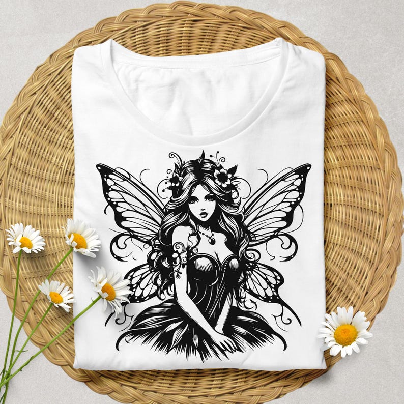 May include: White t-shirt featuring a black and white illustration of a fairy with butterfly wings, long flowing hair, and a floral crown. The shirt is displayed on a woven wicker basket with daisies.