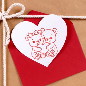 Puede incluir: Una tarjeta blanca en forma de coraz&oacute;n con el contorno rojo de dos osos de peluche abrazando un coraz&oacute;n, colocada sobre un sobre rojo. El sobre est&aacute; sobre una superficie marr&oacute;n, atado con hilo blanco.