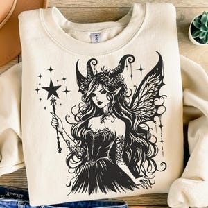 May include: Cream-colored sweatshirt featuring a black graphic of a fairy with long hair, wings, and a star-topped wand. The fairy is adorned with a crown and a dark dress. Stars and decorative lines surround the fairy.