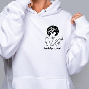 May include: White hoodie with a black and white illustration of a woman with an afro reading a book. The text "Knowledge is power" is printed below the illustration.