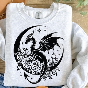 Puede incluir: Una sudadera gris con un dise&ntilde;o gr&aacute;fico en blanco y negro de un drag&oacute;n enroscado alrededor de una luna creciente con rosas y cristales.