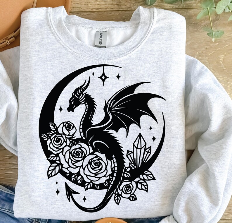 Puede incluir: Una sudadera gris con un dise&ntilde;o gr&aacute;fico en blanco y negro de un drag&oacute;n enroscado alrededor de una luna creciente con rosas y cristales.