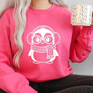 K&ouml;nnte beinhalten: Ein leuchtend pinkfarbenes Sweatshirt mit einem wei&szlig;en Pinguin-Motiv, das Ohrensch&uuml;tzer, eine Brille und einen Schal tr&auml;gt. Das Pinguin-Design befindet sich mittig auf der Vorderseite des Sweatshirts. Die Person h&auml;lt einen wei&szlig;en Becher mit goldenen vertikalen Linien.