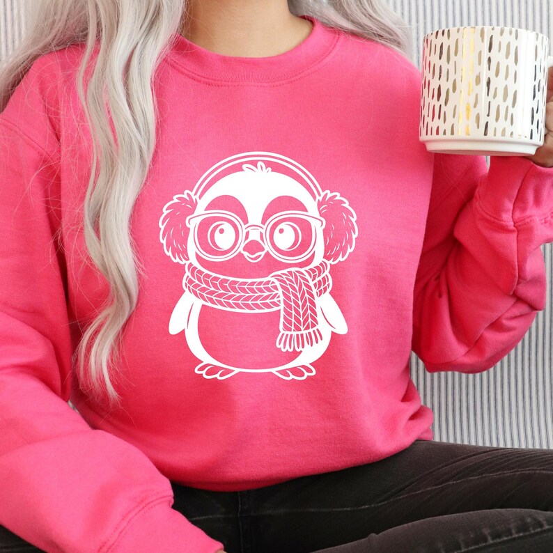 K&ouml;nnte beinhalten: Ein leuchtend pinkfarbenes Sweatshirt mit einem wei&szlig;en Pinguin-Motiv, das Ohrensch&uuml;tzer, eine Brille und einen Schal tr&auml;gt. Das Pinguin-Design befindet sich mittig auf der Vorderseite des Sweatshirts. Die Person h&auml;lt einen wei&szlig;en Becher mit goldenen vertikalen Linien.
