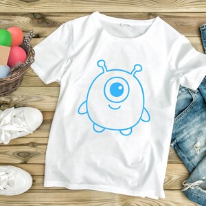 May include: White t-shirt featuring a blue cartoon alien with one eye, antennae, and small arms and legs. The graphic is centered on the shirt. The shirt is displayed on a wooden surface with a pair of white sneakers and a basket of colorful eggs.