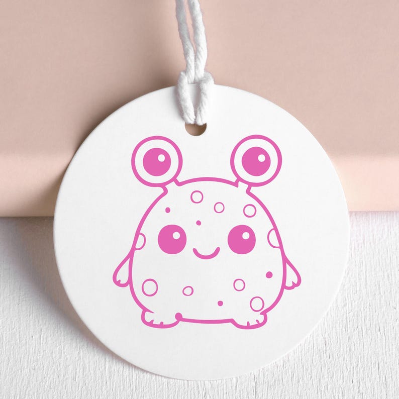 May include: A white, round tag with a pink cartoon alien design. The alien has large eyes, a smiling mouth, and spots. A white string is attached at the top for hanging. The design is simple and playful.
