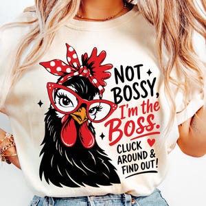 Puede incluir: Camiseta color crema con una ilustración de un pollo negro con gafas rojas y una diadema de lunares. El texto dice "NOT BOSSY, I'm the Boss. CLUCK AROUND & FIND OUT!" en rojo y negro.