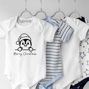 K&ouml;nnte beinhalten: Wei&szlig;e Baby-Bodys, aufgeh&auml;ngt an grauen Kleiderb&uuml;geln. Einer zeigt ein Weihnachtspinguin-Design mit dem Text "Merry Christmas". Ein weiterer hat blau-wei&szlig;e Streifen, andere Sternmuster.