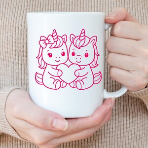 Puede incluir: Taza de cer&aacute;mica blanca con una ilustraci&oacute;n rosa de dos unicornios de dibujos animados sosteniendo un coraz&oacute;n. Un unicornio tiene un lazo, y ambos tienen cuernos y crines rosas. La taza es sostenida por una persona.