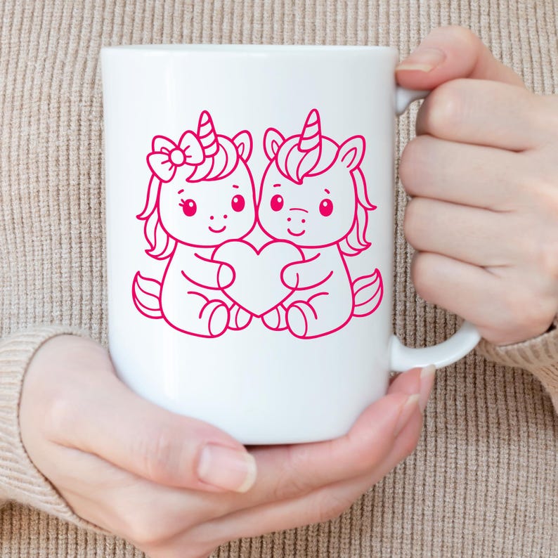 Puede incluir: Taza de cer&aacute;mica blanca con una ilustraci&oacute;n rosa de dos unicornios de dibujos animados sosteniendo un coraz&oacute;n. Un unicornio tiene un lazo, y ambos tienen cuernos y crines rosas. La taza es sostenida por una persona.