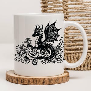 Puede incluir: Taza de cer&aacute;mica blanca con una silueta negra de un drag&oacute;n rodeado de rosas. El drag&oacute;n est&aacute; detallado con intrincados patrones y sostiene una rosa en su boca.