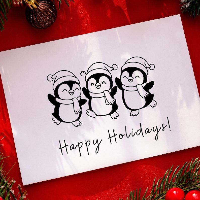 May include: A white greeting card with three cartoon penguins wearing Santa hats and scarves. The penguins are black and white with smiling faces. The words "Happy Holidays!" are written in black script below the penguins. The card is on a red surface.