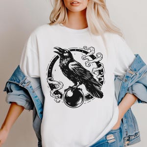 Pu&ograve; includere: T-shirt bianco con un'illustrazione nera di un corvo appollaiato su una sfera di cristallo. Il corvo &egrave; all'interno di una cornice circolare con fumo vorticoso. Il disegno &egrave; centrato sulla parte anteriore della maglietta.