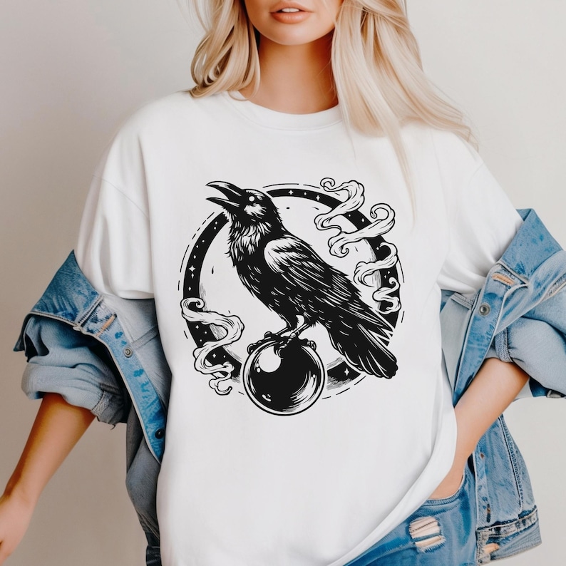 Pu&ograve; includere: T-shirt bianco con un'illustrazione nera di un corvo appollaiato su una sfera di cristallo. Il corvo &egrave; all'interno di una cornice circolare con fumo vorticoso. Il disegno &egrave; centrato sulla parte anteriore della maglietta.
