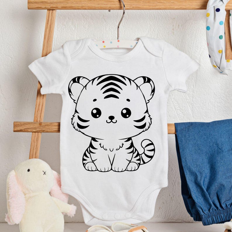 K&ouml;nnte beinhalten: Wei&szlig;er Baby-Body mit einem schwarz-wei&szlig;en Cartoon-Tiger-Design. Der Tiger hat gro&szlig;e Augen und Streifen. Der Body h&auml;ngt an einem Holzb&uuml;gel. Blaue Jeanshose und ein Stoffhase sind im Hintergrund.