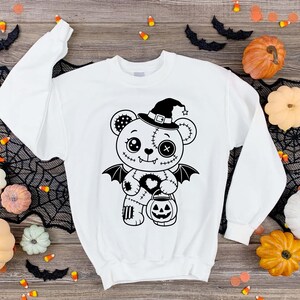 May include: A white long-sleeve sweatshirt featuring a black graphic print of a stitched teddy bear wearing a witch hat and bat wings. The bear holds a jack-o'-lantern bucket, with one button eye, one circular eye, and a heart-shaped patch on its chest.