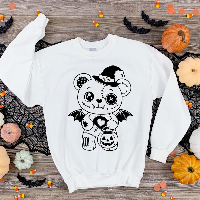 May include: A white long-sleeve sweatshirt featuring a black graphic print of a stitched teddy bear wearing a witch hat and bat wings. The bear holds a jack-o'-lantern bucket, with one button eye, one circular eye, and a heart-shaped patch on its chest.