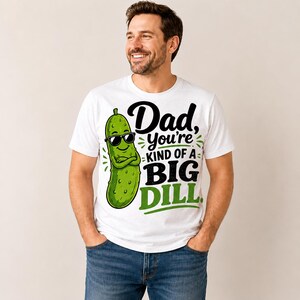 May include: White t-shirt with a graphic of a green pickle wearing sunglasses and the text "Dad, you're kind of a Big Dill." The shirt is a crew neck and is being worn by a person.