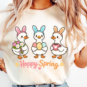 May include: Cream-colored t-shirt featuring three cartoon ducks wearing bunny ears. One duck holds flowers, another holds Easter eggs, and the third holds a mug. The text "Hoppy Spring" is below the ducks, with floral accents.