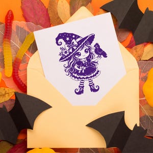 May include: A white greeting card featuring a purple ink illustration of a character wearing a witch hat and dress, holding a small bird. The card is tucked into a light yellow envelope, surrounded by black paper bats and colorful gummy worms.