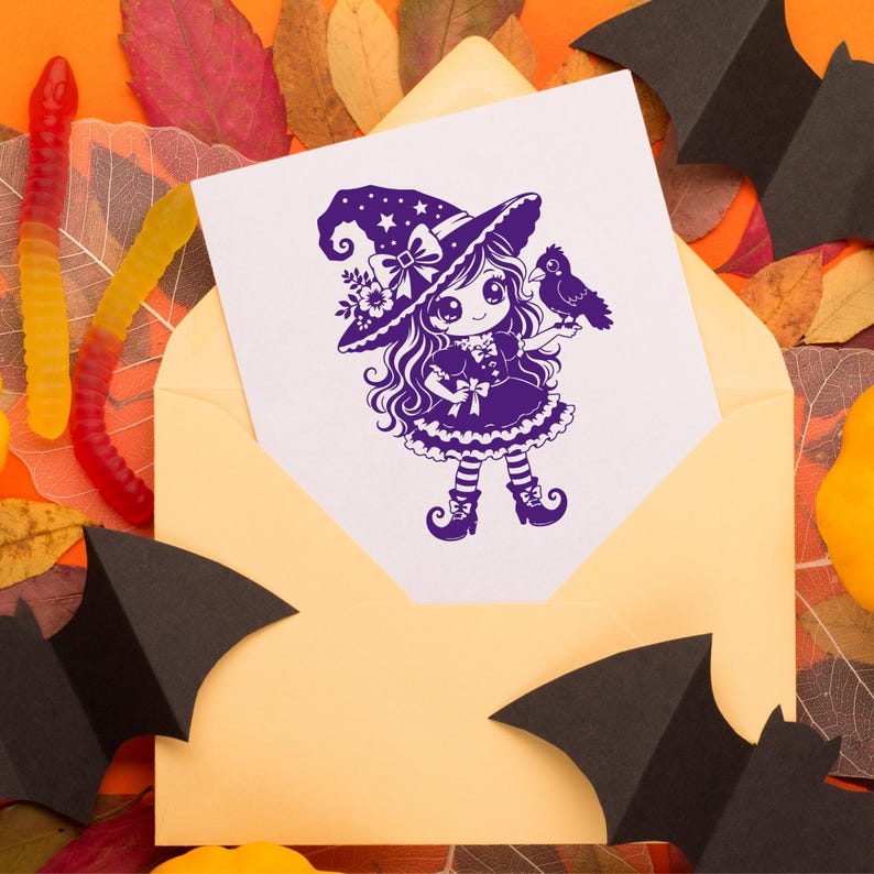 May include: A white greeting card featuring a purple ink illustration of a character wearing a witch hat and dress, holding a small bird. The card is tucked into a light yellow envelope, surrounded by black paper bats and colorful gummy worms.