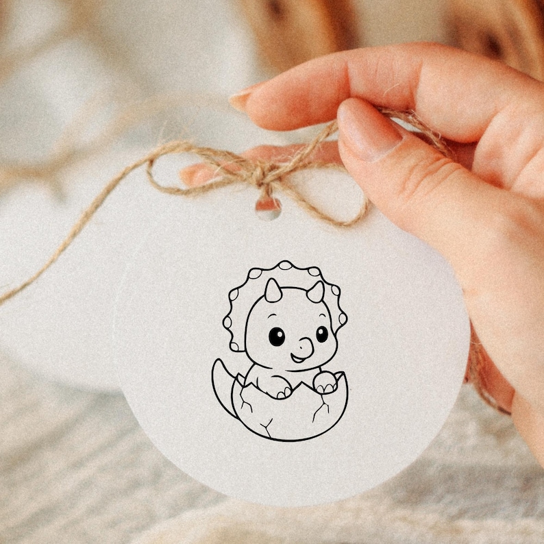 May include: A white, round ornament featuring a black and white illustration of a baby triceratops hatching from an egg. The ornament is strung with twine and has a small hole at the top for hanging.