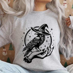 Pu&ograve; includere: Una felpa grigia con una grafica in bianco e nero di un corvo con un cappello da strega, appollaiato su una mano. Il corvo &egrave; circondato da stelle all'interno di una cornice circolare. Il disegno &egrave; stampato su una felpa grigia.