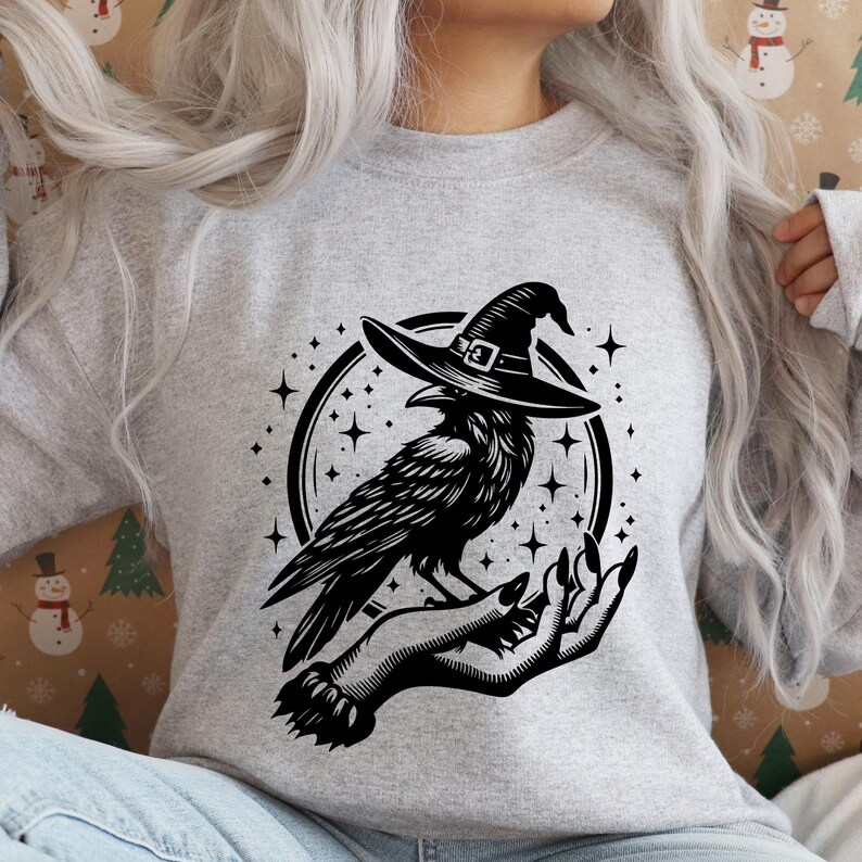Pu&ograve; includere: Una felpa grigia con una grafica in bianco e nero di un corvo con un cappello da strega, appollaiato su una mano. Il corvo &egrave; circondato da stelle all'interno di una cornice circolare. Il disegno &egrave; stampato su una felpa grigia.
