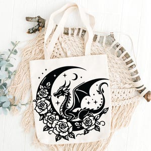 Puede incluir: Una bolsa de lona blanca con un dise&ntilde;o de silueta negra de un drag&oacute;n enroscado alrededor de una luna creciente con estrellas y rosas.