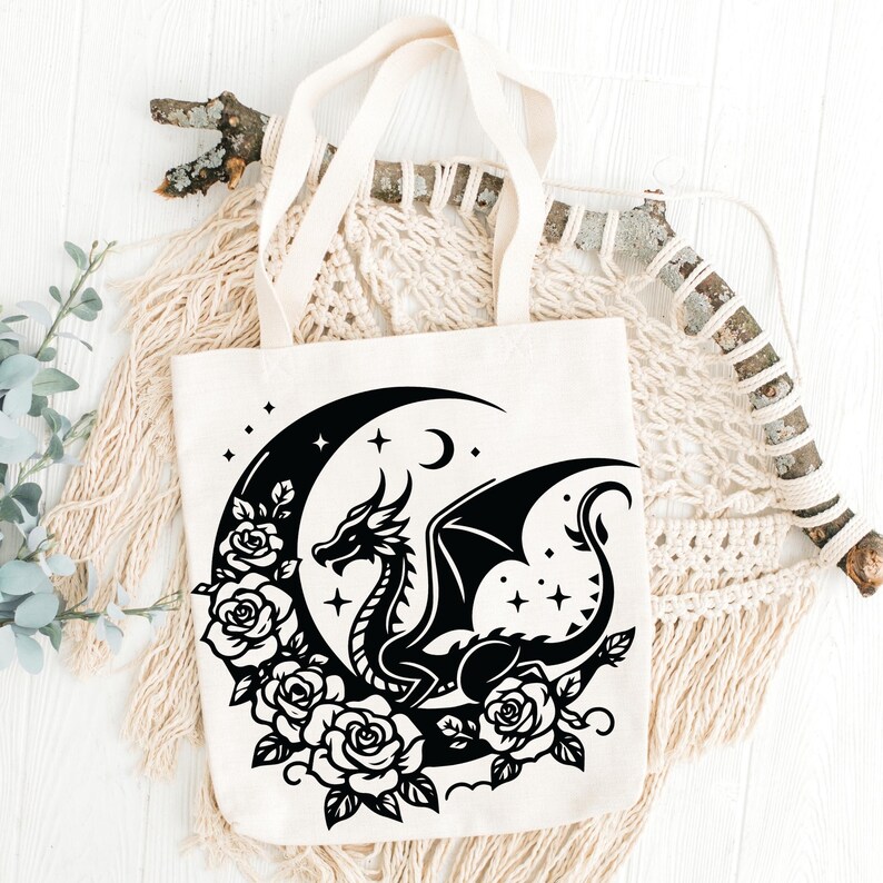 Puede incluir: Una bolsa de lona blanca con un dise&ntilde;o de silueta negra de un drag&oacute;n enroscado alrededor de una luna creciente con estrellas y rosas.